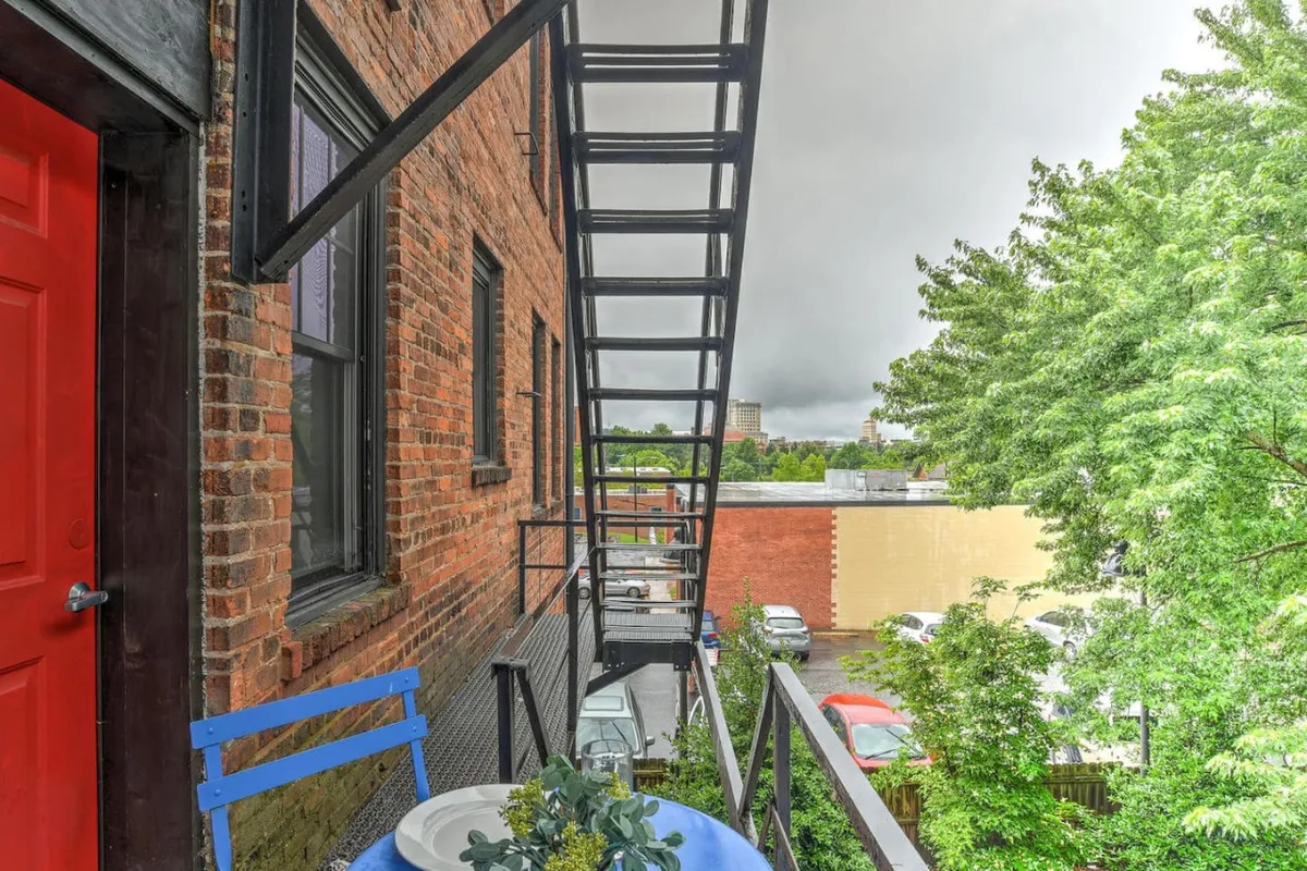 This exterior shot focuses on the building's welcoming red door framed by greenery, with a metal fire escape and a blue chair on the balcony. The casual scene shows the possibility of enjoying coffee outdoors while observing the city, reinforcing the building’s inviting and lively surroundings.