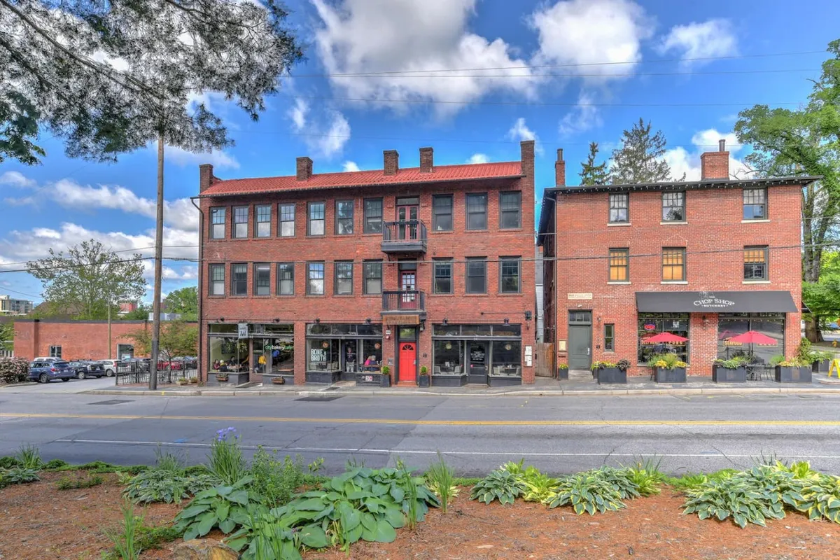 A warmly inviting view of the building’s historic brick architecture featuring red doors, an outdoor seating area, and lush landscaping. The scene emphasizes the welcoming ambiance of the entry and hints at the building’s storied past, aligning with the narrative of an inviting and historic entryway.