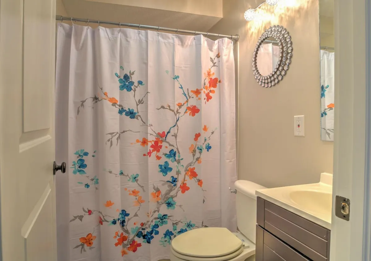 This image features a freshly renovated bathroom with a modern vanity, mirrored decor, and a stylish floral shower curtain. The scene feels calm and refreshing, providing a sense of privacy and comfort that complements the adjacent bedroom.