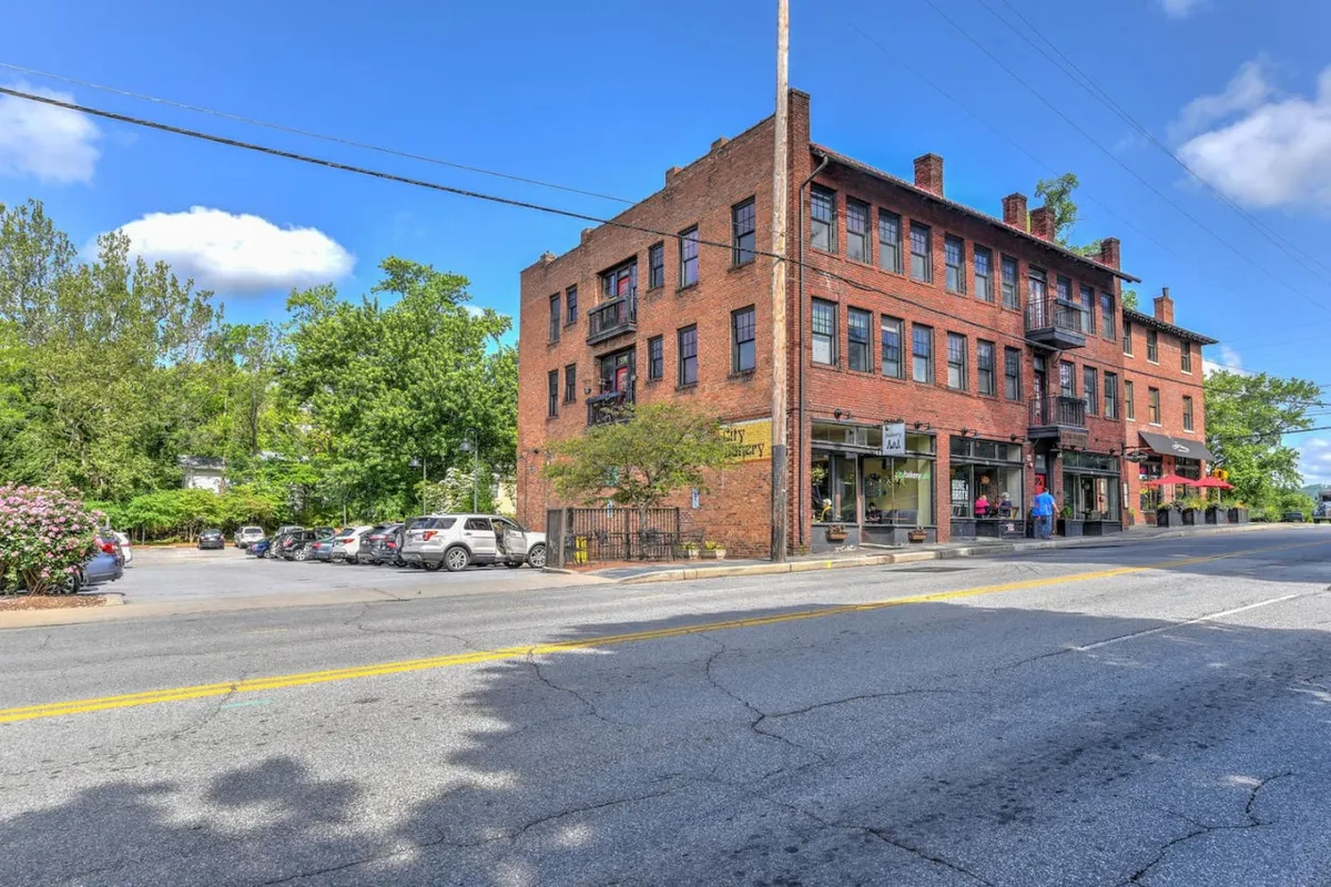 This exterior image showcases the brick facade of the King James Building, with street-level storefronts, leafy trees, and parked cars creating a lively urban atmosphere. The building’s historic character is evident in its architectural details, providing immediate context for the property's location in North Asheville.