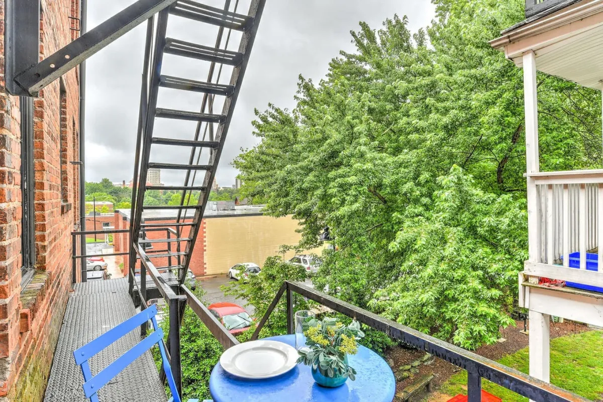 This exterior image captures a serene balcony scene with a blue table nestled among lush green foliage and set against the backdrop of a classic fire escape staircase. The mood is tranquil and cozy, suggesting an inviting space for outdoor dining or quiet moments enjoying nature. The ambiance evokes the historical charm and peace described in the section.