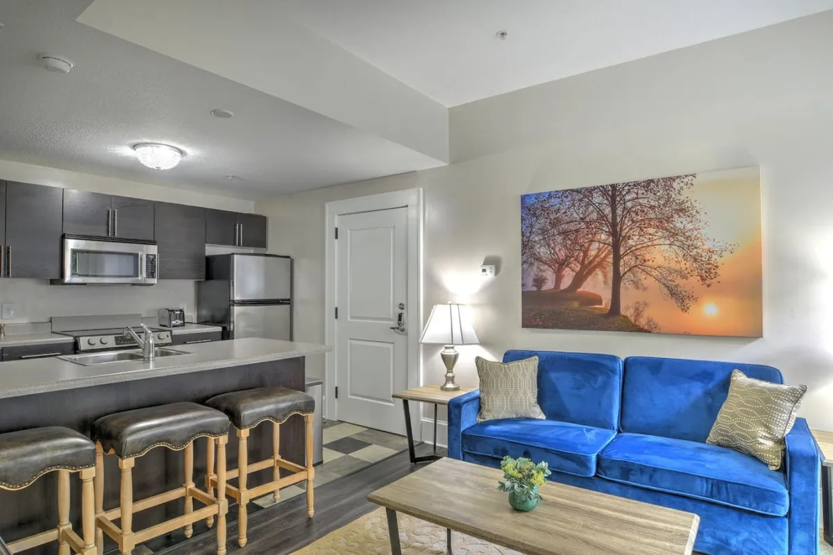 A view of the living area highlighting the blue couch, an adjacent kitchen bar, and stylish lighting. Sunlight streams in through the windows, casting a warm glow across the room. The open layout suggests versatility for both relaxing and working, with a table lamp hinting at a potential workspace nook.