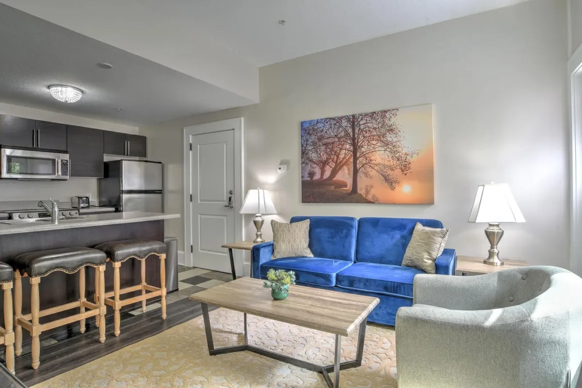 A cozy living space featuring a plush blue sofa paired with a wooden coffee table, set against a backdrop of large windows that allow natural sunlight to flood the room. The kitchenette is visible in the background, and art adorns the walls, adding a touch of personality and comfort. The layout allows for both relaxation and casual gatherings.