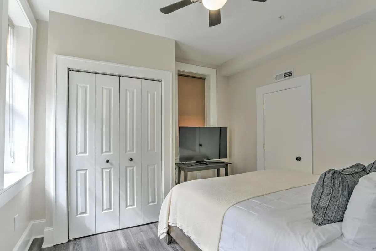 This image shows a cozy bedroom featuring a plush bed dressed in crisp white linens, a television mounted opposite the bed, and closet doors suggesting storage space. The soft lighting and inviting setup evoke comfort and relaxation, perfectly suited for restful nights or leisurely mornings.